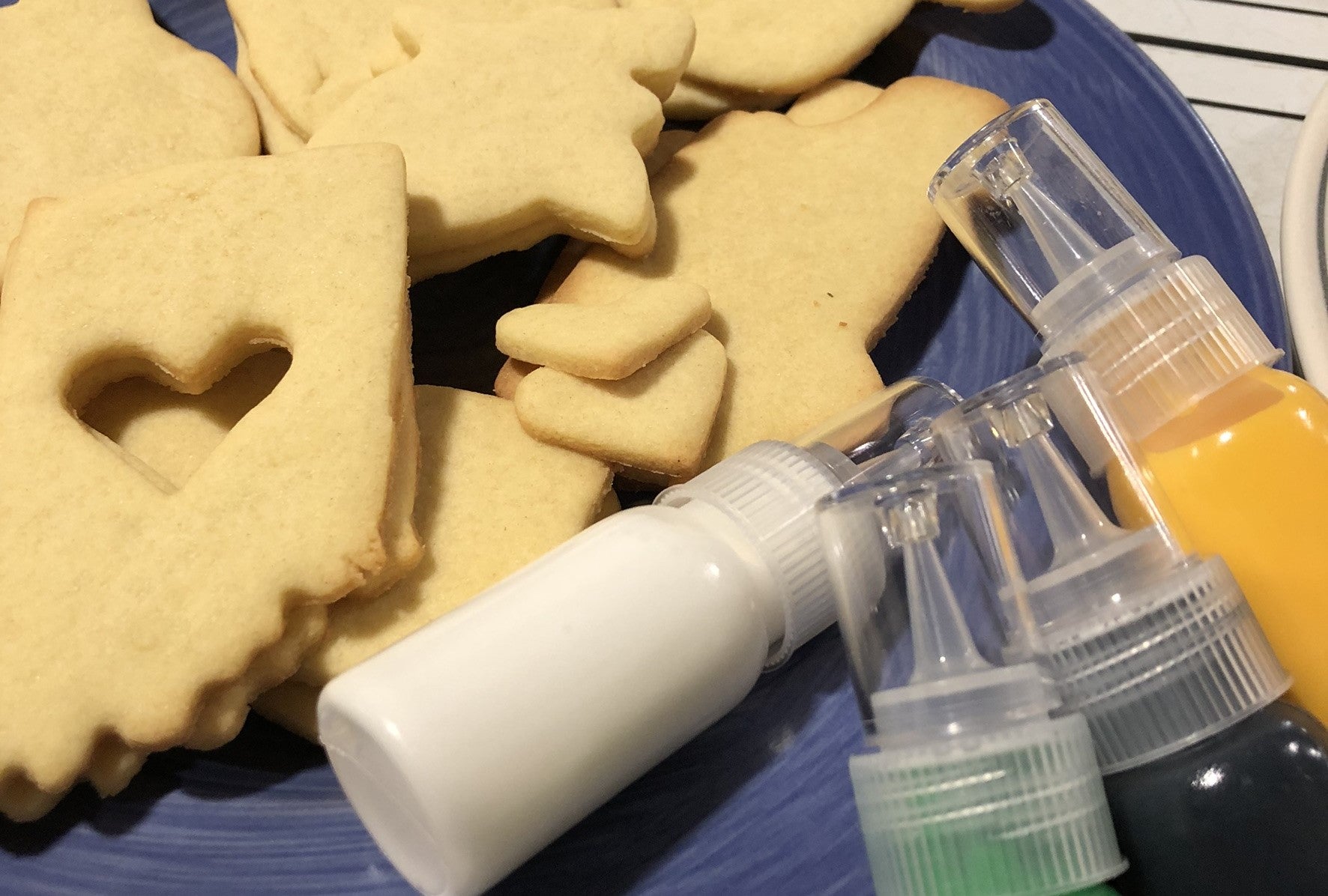 Freshly baked sugar cookies in various shapes, displayed on a blue plate with colorful icing bottles ready for decorating.